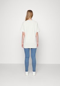 Camiseta gris claro de manga corta, ajuste relajado, con un dobladillo trasero ligeramente más largo, combinada con jeans ajustados azules y zapatillas blancas.