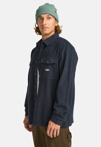 Marineblauwe langemouwen shirt met knoopsluiting, twee borstzakken en een geborduurd logo. Katoenen materiaal met een gladde textuur.