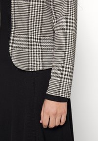 Svartvitt houndstooth-blazer med en figursydd form och textilstruktur, tillsammans med en svart ribbad klänning. Har en slät krage och manschetter.