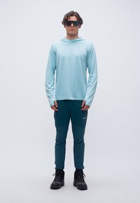 Άνδρας που στέκεται, φοράει ανοιχτό μπλε μακρυμάνικη φούτερ με κουκούλα, σκούρο μπλε παντελόνι, μαύρα παπούτσια πεζοπορίας και μεγάλα αντανακλαστικά γυαλιά ηλίου.