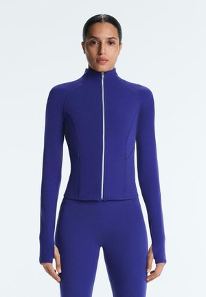 Žena stojaca čelom k fotoaparátu, oblečená v priliehavom fialovom farebnom zipsovom bundu s dlhými rukávmi a zodpovedajúcom legínach na jednoduchom svetlom pozadí.