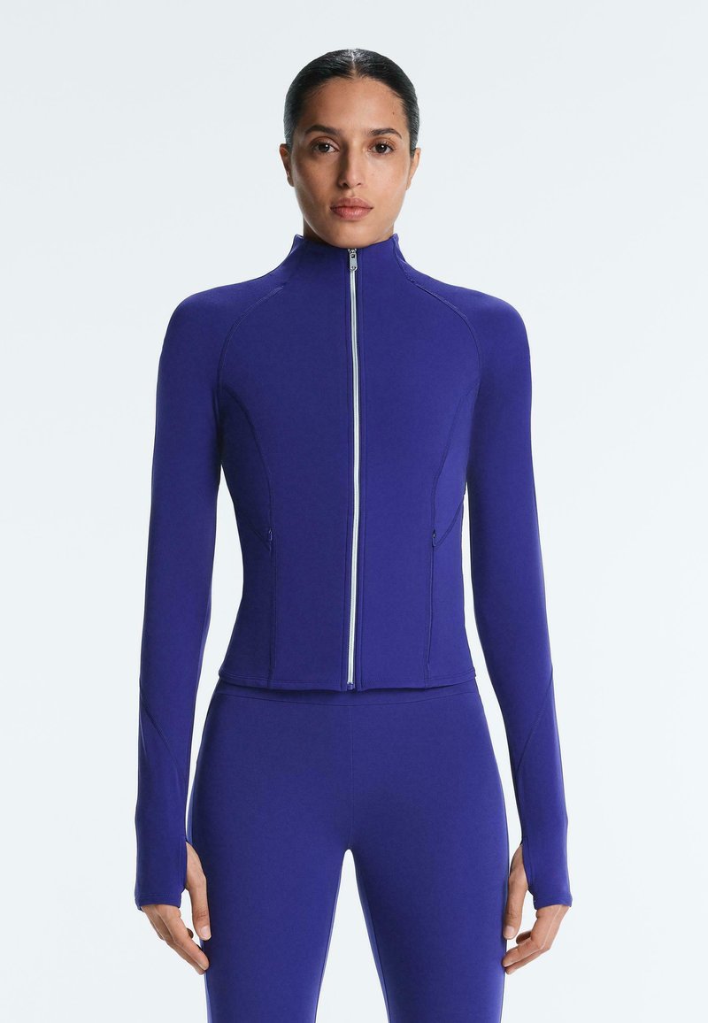 Femme debout face à l'avant, portant une veste zippée moulante à manches longues violette et un legging assorti sur un fond clair uni.