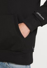 Felpa nera con polsini a costine, tasca a marsupio e discreto dettaglio del logo "Calvin Klein" sulla manica. Miscela di cotone, texture morbida.