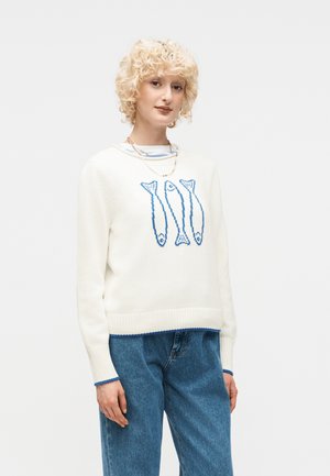 Młoda osoba o kręconych blond włosach nosi biały sweter z wzorem trzech niebieskich ryb oraz niebieskie dżinsy na jednolitym tle.