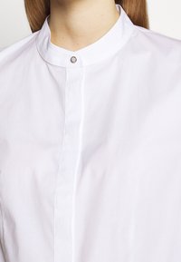Camicia bianca con colletto realizzata in tessuto morbido, con chiusura a bottoni al collo e un sottile design della pattina verticale.