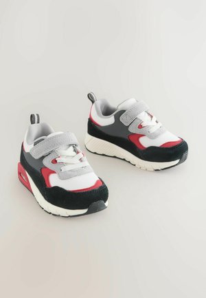 Baltas sietveida sporta apavi ar pelēkiem, sarkaniem un melniem akcentiem. Iekļauj Velcro siksnu, auklu detaļas un teksturētu gumijas zoli.