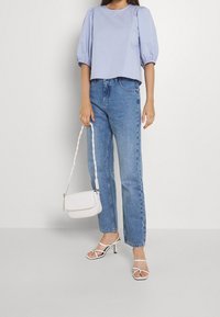 Kvinna i en ljusblå puffärmad topp, högmidjade blå jeans, vita remsandaler samt hållande en vit axelväska.