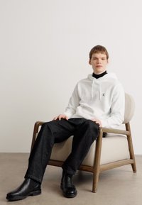 Sudadera blanca con logo, cuello alto negro, jeans oscuros y botas chelsea negras. Sentado en una silla de color crema claro con patas de madera frente a un fondo liso.