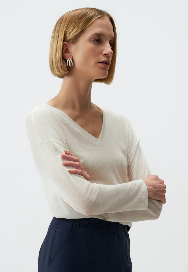 V-hals lange mouwen top in lichtgewicht, doorschijnende crèmewitte stof. Gecombineerd met donkerblauwe broek. Model heeft een kort kapsel en zilveren oorringen.