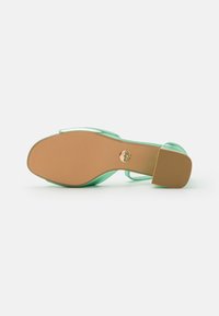 Transparent högklackad sandal i mintgrönt med en beige gummisula och en rund klack som har en guldfärgad logodetalj.
