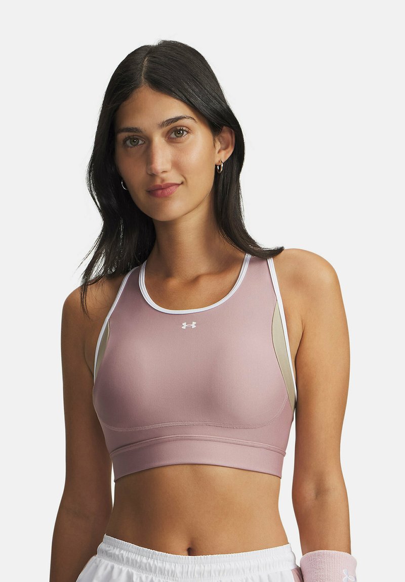Young woman with long dark hair wearing a pink and beige sports bra and white shorts, posing against a plain background.