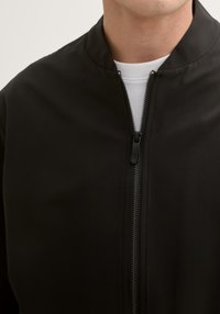 Czarna kurtka bombowa z stójką, pełnym zamkiem z przodu i gładką fakturą materiału. Posiada kontrastujący biały materiał wewnętrzny.