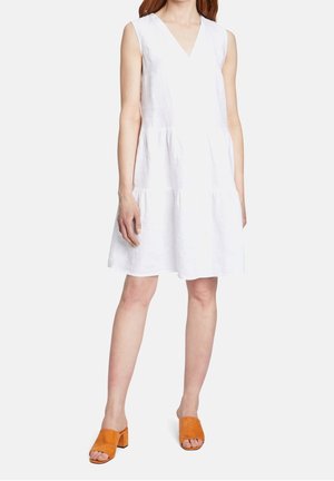 Frau trägt ärmelloses weißes knielanges Kleid mit V-Ausschnitt und orangefarbene Sandalen mit offenem Zehenbereich und Blockabsatz, steht vor einfarbigem Hintergrund.