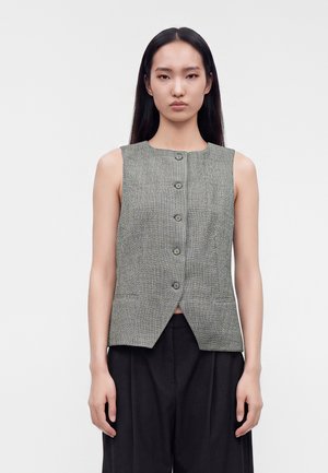 Vrouw met lang zwart haar die een grijze mouwloze gebuttonde vest en zwarte wijde broek draagt, staand tegen een effen lichte achtergrond.