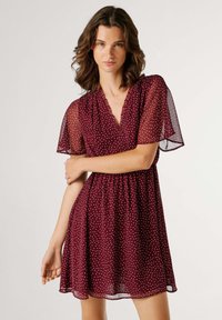 Vestido burdeos con lunares, mangas cortas y transparentes, escote en V y cintura elástica, que se despliega en una falda acampanada.