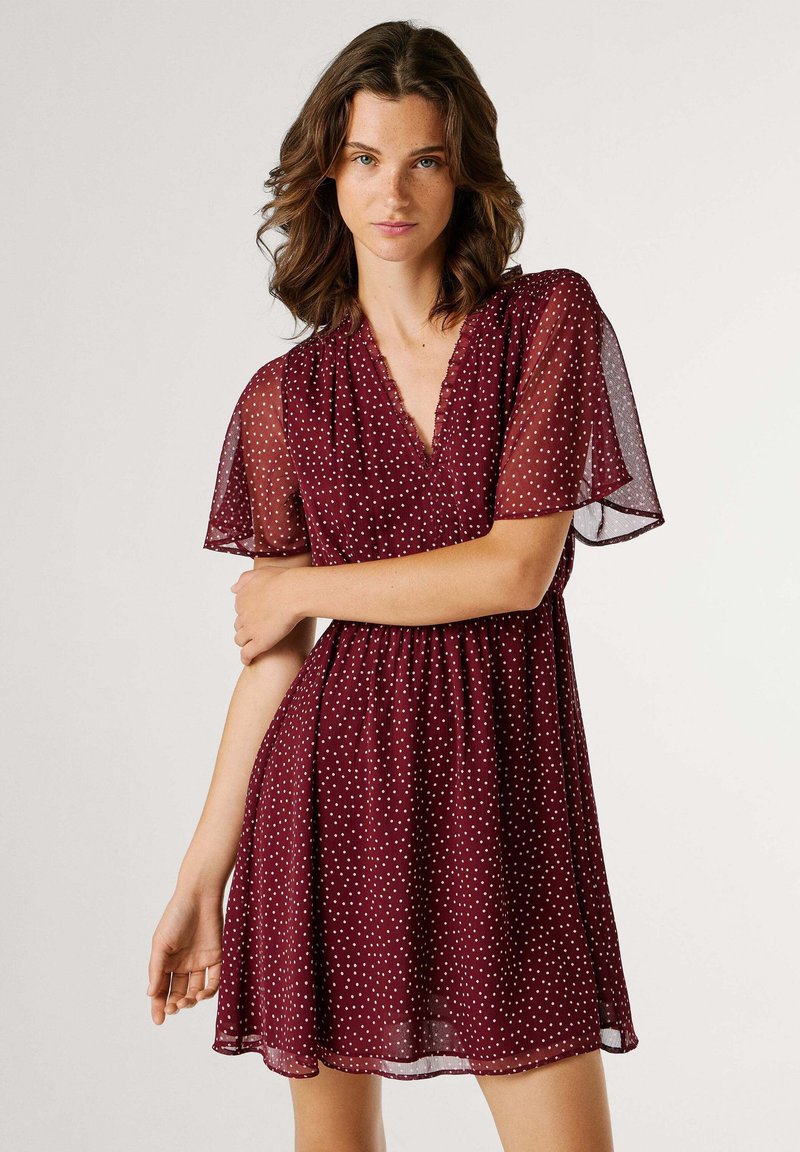 Vestido burdeos con lunares, mangas cortas y transparentes, escote en V y cintura elástica, que se despliega en una falda acampanada.