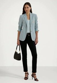 Ženska v svetlo modrem sakou, belem topu, črnih hlačah in črnih sandalah s petami, ki drži majhno črno torbico na belem ozadju.