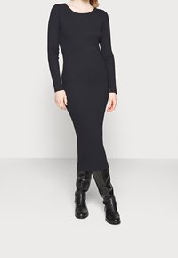 Vestido midi negro de manga larga con cuello redondo, que presenta un corte ajustado y una textura sutil. Combinado con botas negras hasta la rodilla.