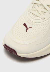 Beige sportssko med en teksturert overdel, som har et bordeauxfarget logo og detaljer, rund tå og perforerte detaljer for ventilasjon.