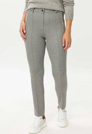 Vrouw draagt grijze slim-fit broek met voornaad, een grijze trui en witte sneakers, staand voor een witte achtergrond.