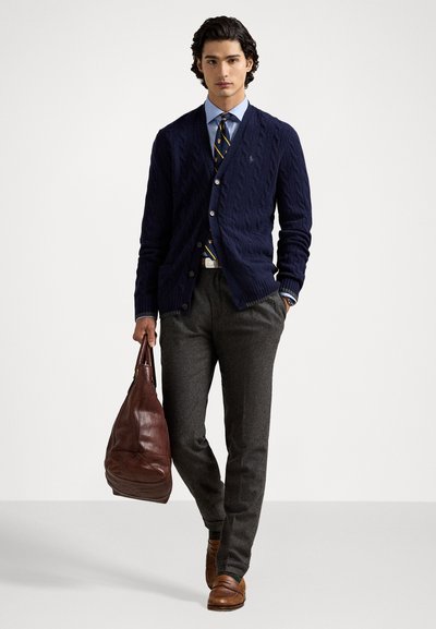 Cardigan en maille torsadée bleu marine pour homme, chemise bleu clair, cravate à motifs, pantalon gris, sac en cuir marron, chaussures marron et une ceinture blanche.