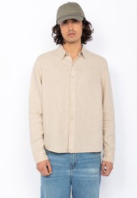 Beige linnen overhemd met lange mouwen en een losse pasvorm, gecombineerd met een lichtgrijze pet. Voorzien van subtiele logo borduursels en knopen.