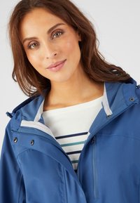 Chaqueta impermeable azul con capucha, que presenta un bolsillo con cremallera, detalles en gris, y una camisa blanca debajo con rayas azules.