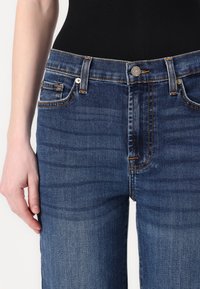 Mørkeblå denimjeans med fem-lommers design, sølvfarget detaljer og oransje sømmer. Tøyet har en svak falmende effekt.