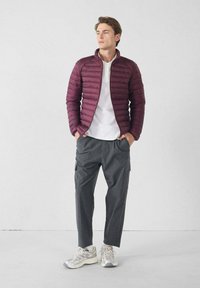 Bordeaux pufferjack met horizontale stiksels, witte polo, donkergrijze cargobroek en grijze sneakers. Casual outfit getoond.