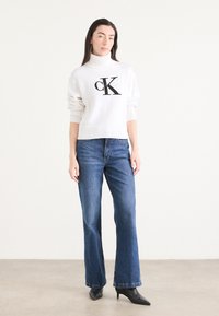 Hvit genser med høy hals og svart "CK" logo, kombinert med blå rette jeans og spisse svarte hæler, satt mot en nøytral bakgrunn.