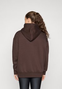 Overdimensjonert brun hettegensere med stor hette, ribbede mansjetter og nederkant. Glatt tekstur og avslappet passform. Brukt med svarte leggings.
