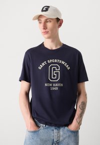 Mørkeblå bomullst-skjorte med rund hals, som har et kremfarget grafisk logo med teksten "GANT SPORTSWEAR NEW HAVEN 1949." Brukt med slitte denim.