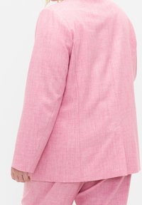 Personne portant une veste de costume rose texturée et un pantalon assorti, vue de dos et de côté, avec une coupe décontractée.