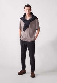 Camisetta a maniche corte a coste grigio chiaro, maglione nero a girocollo appoggiato sulle spalle, abbinato a pantaloni neri e scarpe in suede marroni.