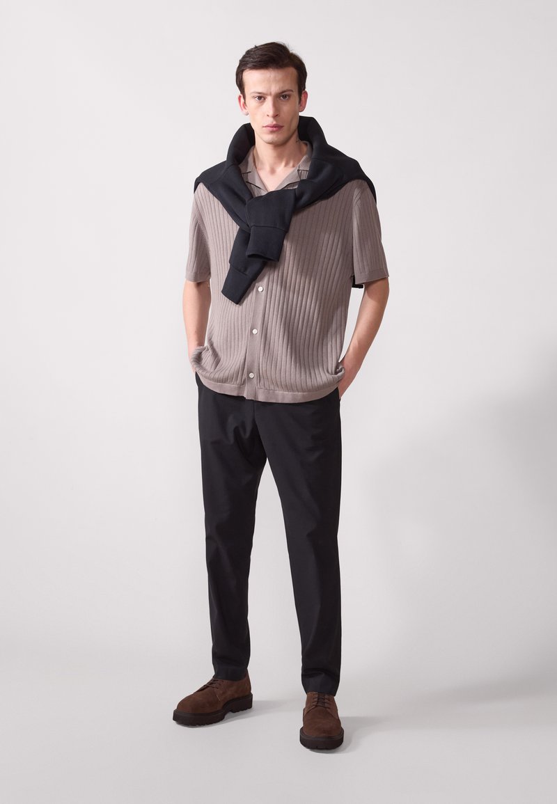 Camisetta a maniche corte a coste grigio chiaro, maglione nero a girocollo appoggiato sulle spalle, abbinato a pantaloni neri e scarpe in suede marroni.