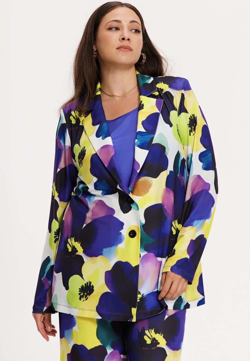 Femme portant un haut violet avec un blazer assorti et un pantalon ornés de grands motifs floraux jaunes, bleus et violets, debout devant un fond blanc.