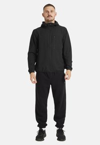Sort sportsjakke med lynlås foran, elastiske ærmer og hætte med snøre, sat sammen med sorte joggers og sorte sneakers. Glat tekstur.