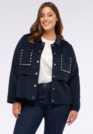 Veste marine en tissu doux avec des accessoires en or, dotée d'accents cloutés, de deux poches avant et d'un col.