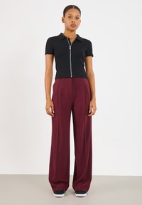 Top preto de manga curta com fecho, combinado com calças largas de cintura alta em burgundy, e sapatilhas pretas e brancas, contra um fundo neutro.