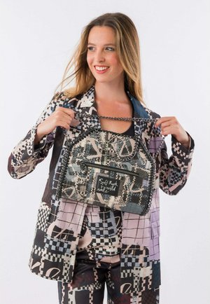 Giovane donna sorridente, che tiene una borsa a tracolla con catena e motivo, indossa un completo abbinato con stampa geometrica astratta in tonalità tenui.