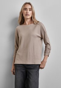 Top beige a maniche lunghe con una texture a costine, righe diagonali e dettagli delle maniche arricciate, abbinato a jeans scuri.