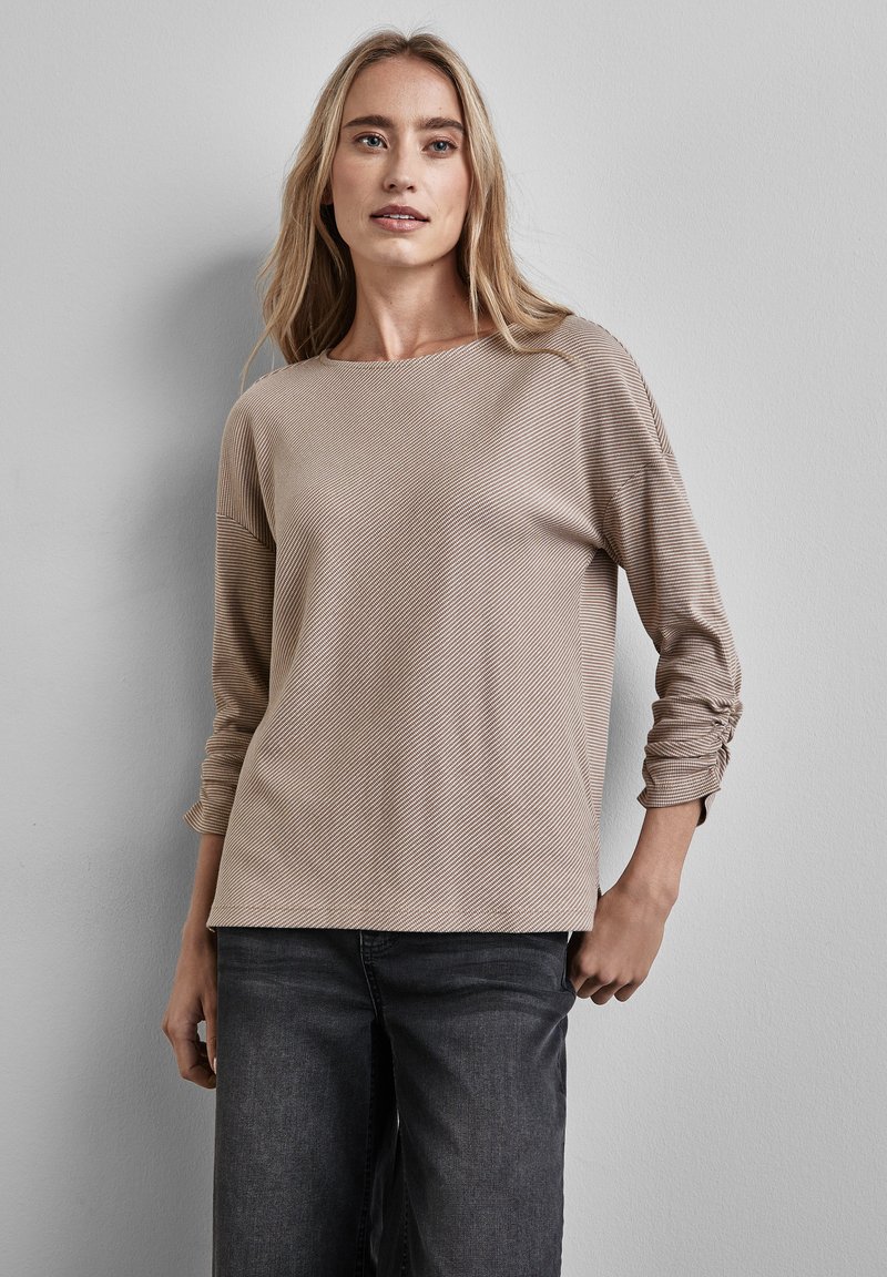 Top beige a maniche lunghe con una texture a costine, righe diagonali e dettagli delle maniche arricciate, abbinato a jeans scuri.