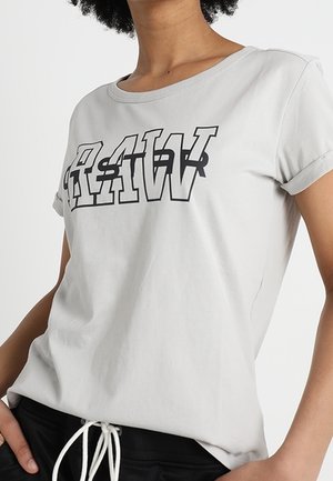 Woman wearing white t-shirt with rolled sleeves and black graphic text "RAW" partially obscured by a black line across the chest.