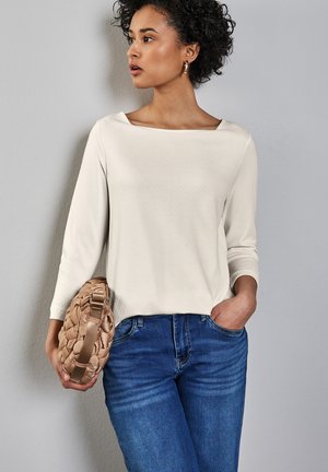 Haut beige clair à manches longues avec un décolleté carré, associé à un jean bleu moyen. Un sac à main tissé marron clair est tenu dans une main.