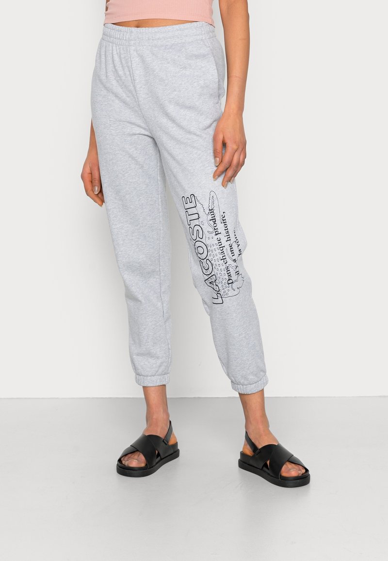 Femme portant un pantalon de survêtement Lacoste gris clair avec un logo noir sur la jambe gauche et des sandales noires à bouts ouverts, debout sur un sol uni.