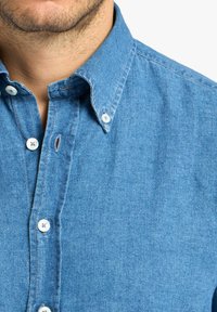 Camicia di jeans blu con colletto a punta, bottoni bianchi e cuciture in evidenza. La trama del tessuto è morbida e presenta un aspetto casual.