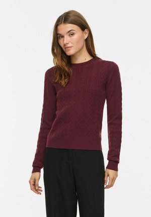 Vrouw met lang bruin haar die een getextureerde bordeauxrode trui en zwarte broek draagt, staand tegen een witte achtergrond.