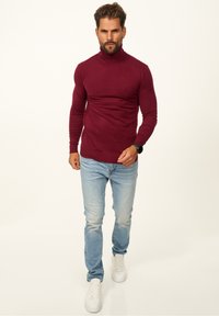Burgunderfarbener Rollkragenpullover, taillierte Passform, gerippte Textur, kombiniert mit ausgebeulten blauen Jeans und weißen Sneakers, die einen lässigen Look erzeugen.