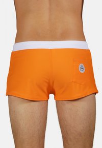 Shorts de bain orange en tissu lisse et extensible. Dotés d'une taille blanche et d'une poche arrière avec un patch logo rond.