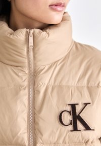 Veste doudoune beige avec un col haut et une fermeture éclair à l'avant. Présente un logo "cK" brodé brun foncé sur le côté gauche. Texture lisse et matelassée.